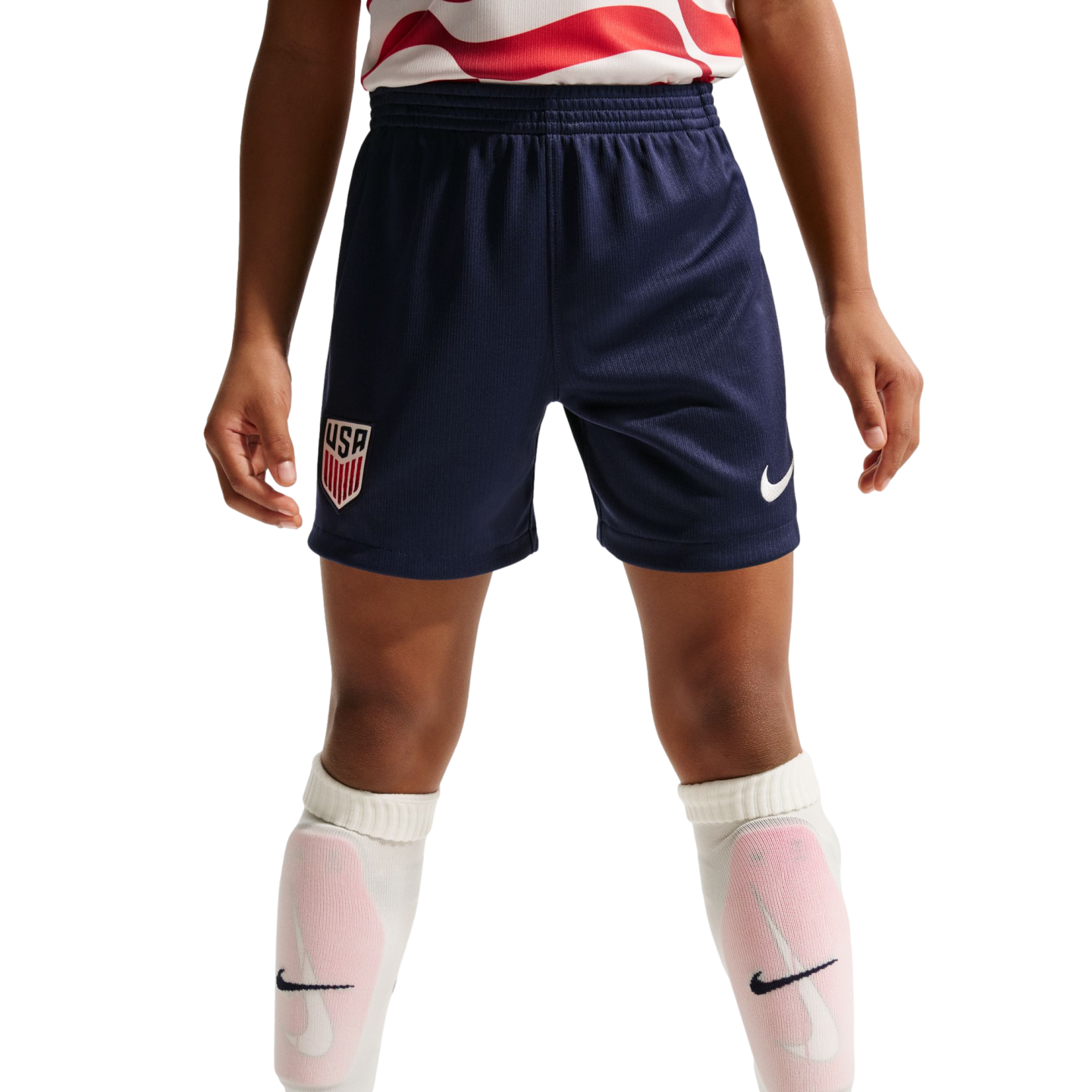 A child wears navy blue USA soccer shorts with a Nike logo, a red and white jersey, and white Nike socks with pink shin guards against a black background.