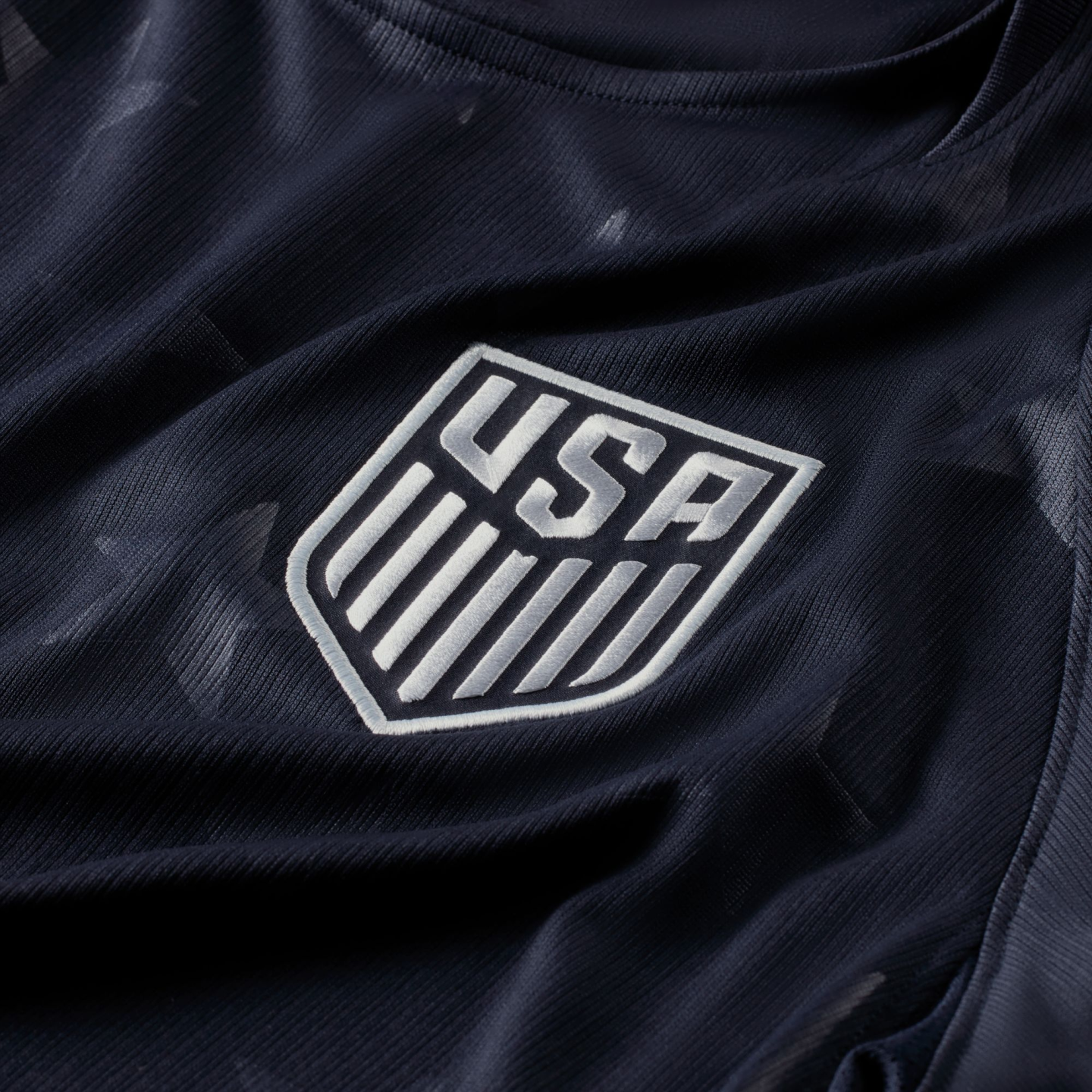 Close-up of a navy blue USA soccer jersey featuring a silver embroidered national team crest and subtle star patterns on the fabric.