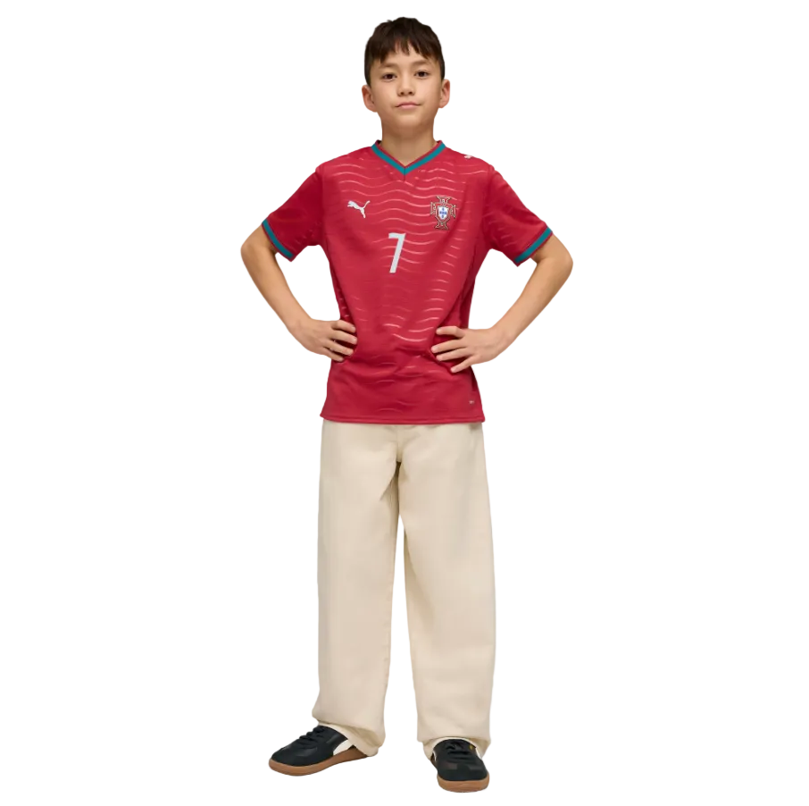 A boy posing in a red soccer jersey with the number 7, wide-leg cream trousers, and black sneakers against a black background.