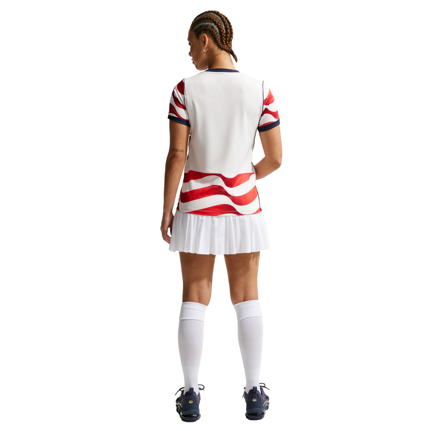 Rear view of a person in a white sports jersey with red wavy stripes, a white pleated skirt, and knee-high socks against a black background.