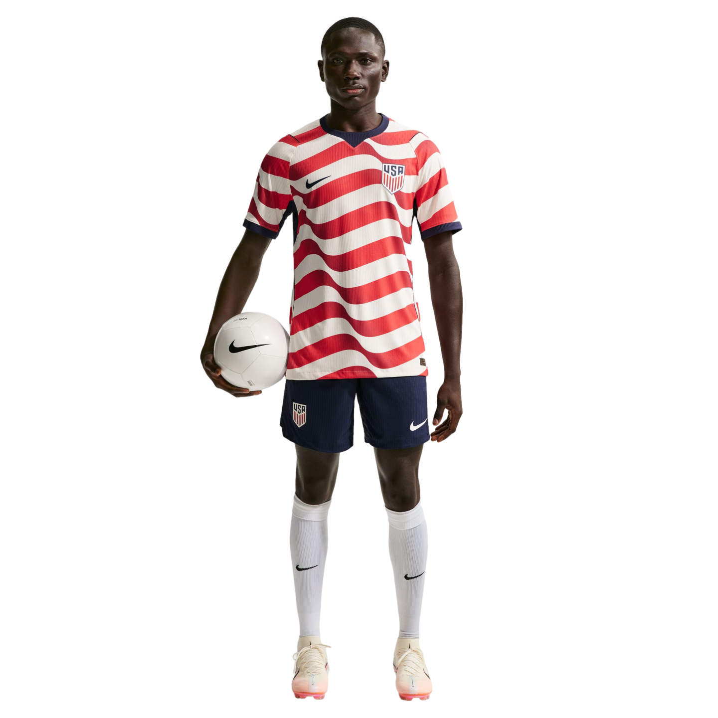 A male soccer player in a wavy red and white USA jersey and navy shorts, holding a soccer ball under his arm against a black background.