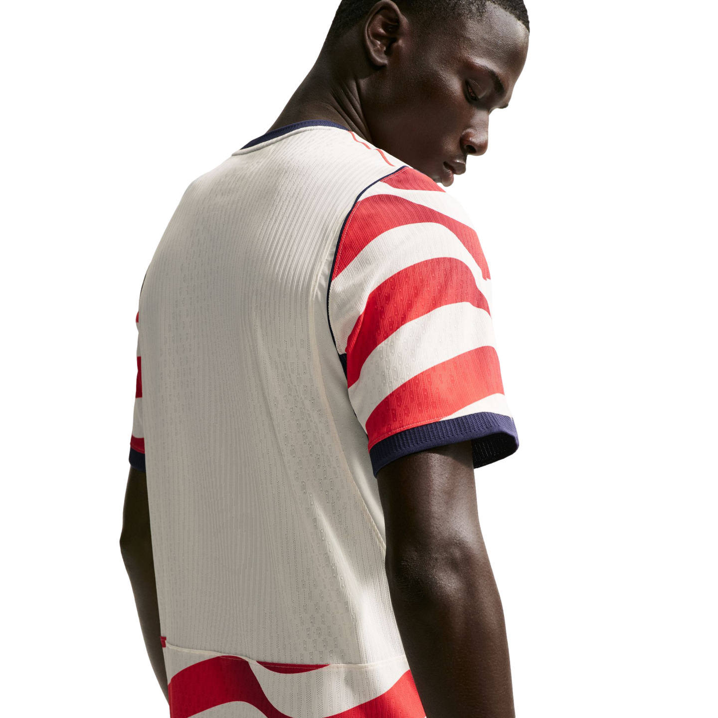 Rear view of a man looking down, wearing a white soccer jersey with red and white striped sleeves and navy blue trim against a black background.