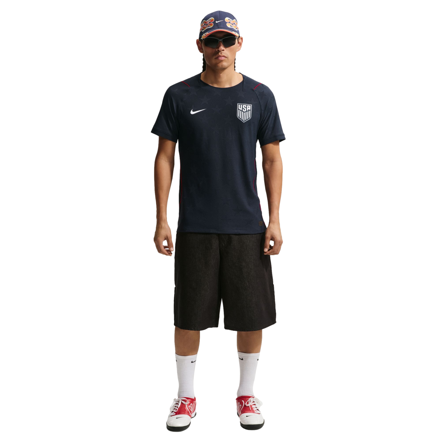 A man in a navy USA soccer jersey, black shorts, and red sneakers stands against a black background wearing a patterned cap and sunglasses.