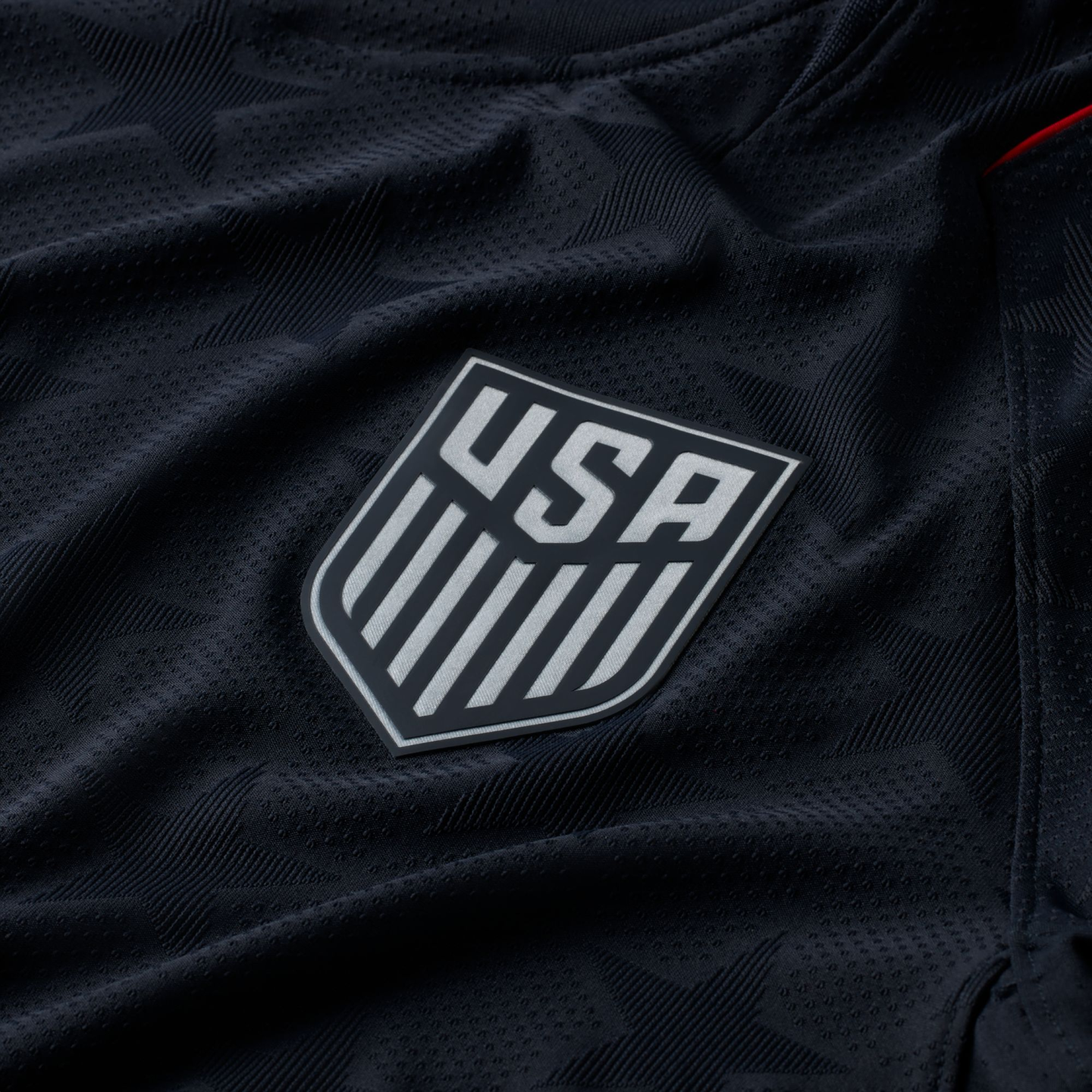 Close-up of a black US Soccer jersey featuring a silver crest and a subtle, tonal star pattern in the textured fabric.