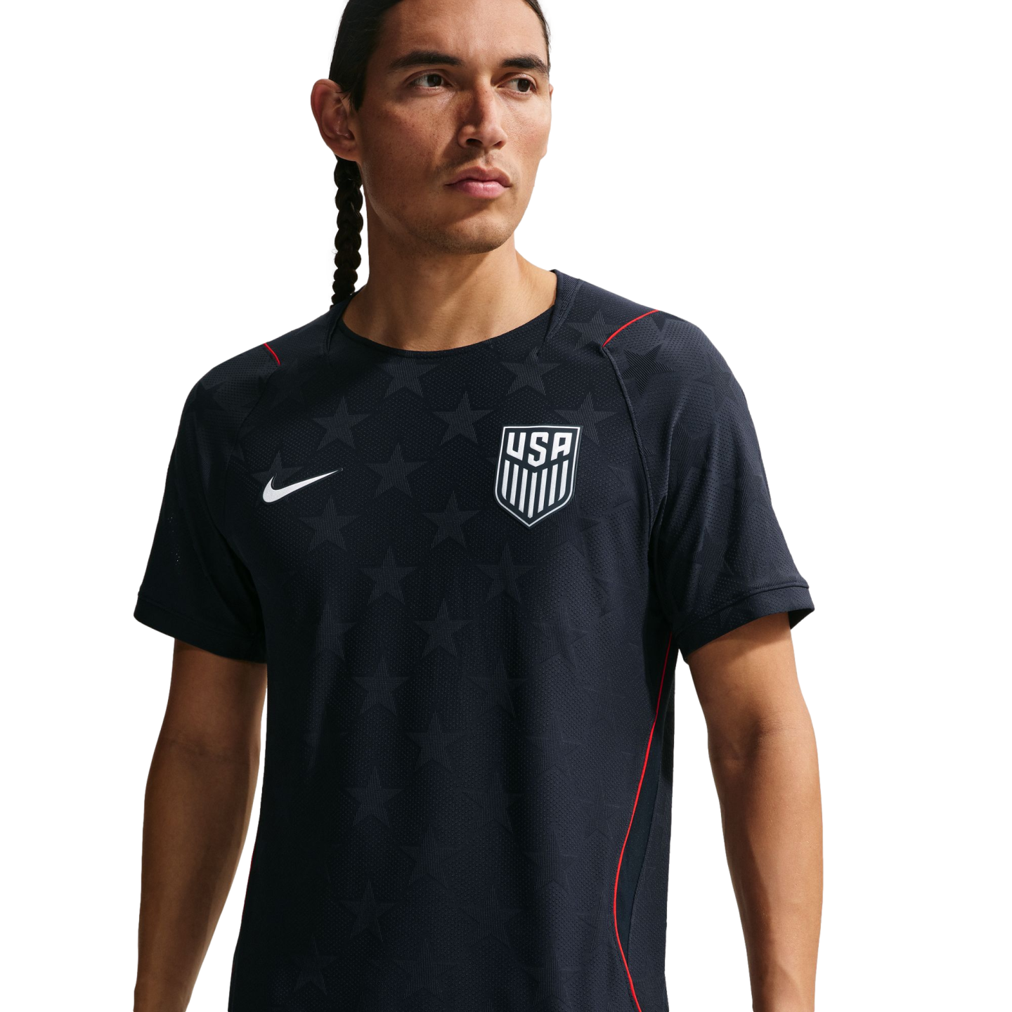 A man wearing a navy blue Nike USA soccer training jersey with star patterns and red accents, looking away from the camera against a black background.