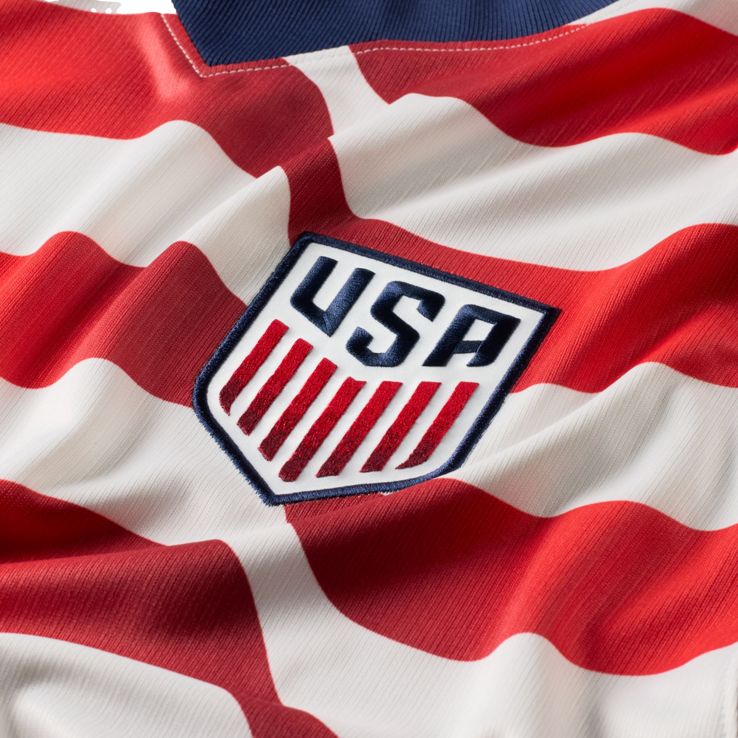 Close-up of a red and white striped soccer jersey with an embroidered shield crest featuring blue "USA" lettering and red vertical stripes.