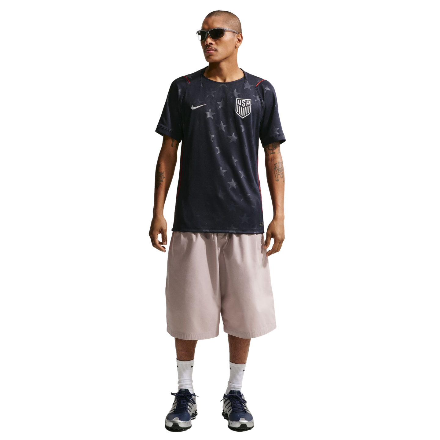 A man models a dark blue Nike USA soccer jersey with grey stars, oversized light pink shorts, and blue sneakers against a black background.