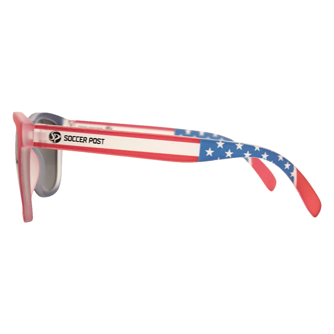 A side view of sunglasses with an American flag design on the temples. The temples feature red and white stripes with a blue, star-patterned ear piece, and the "Soccer Post" logo and name printed on the side. The front frame is red with dark lenses.
